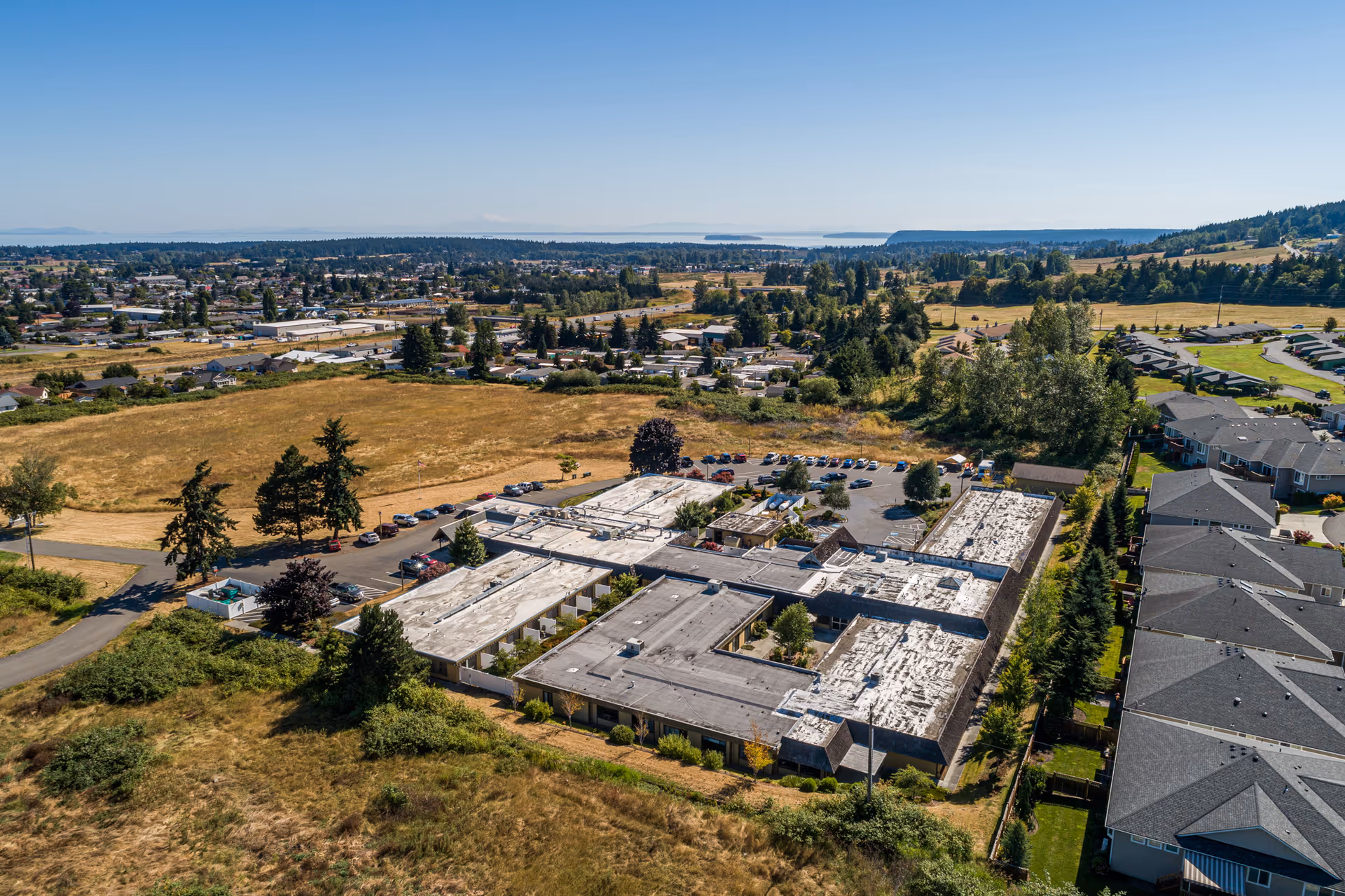 Aerial view of Avamere Olympic Rehabilitation of Sequim, a large single-story building with a flat roof surrounded by parking lots, trees, and open grassy fields. Residential houses and a townscape are visible in the background under a clear blue sky.