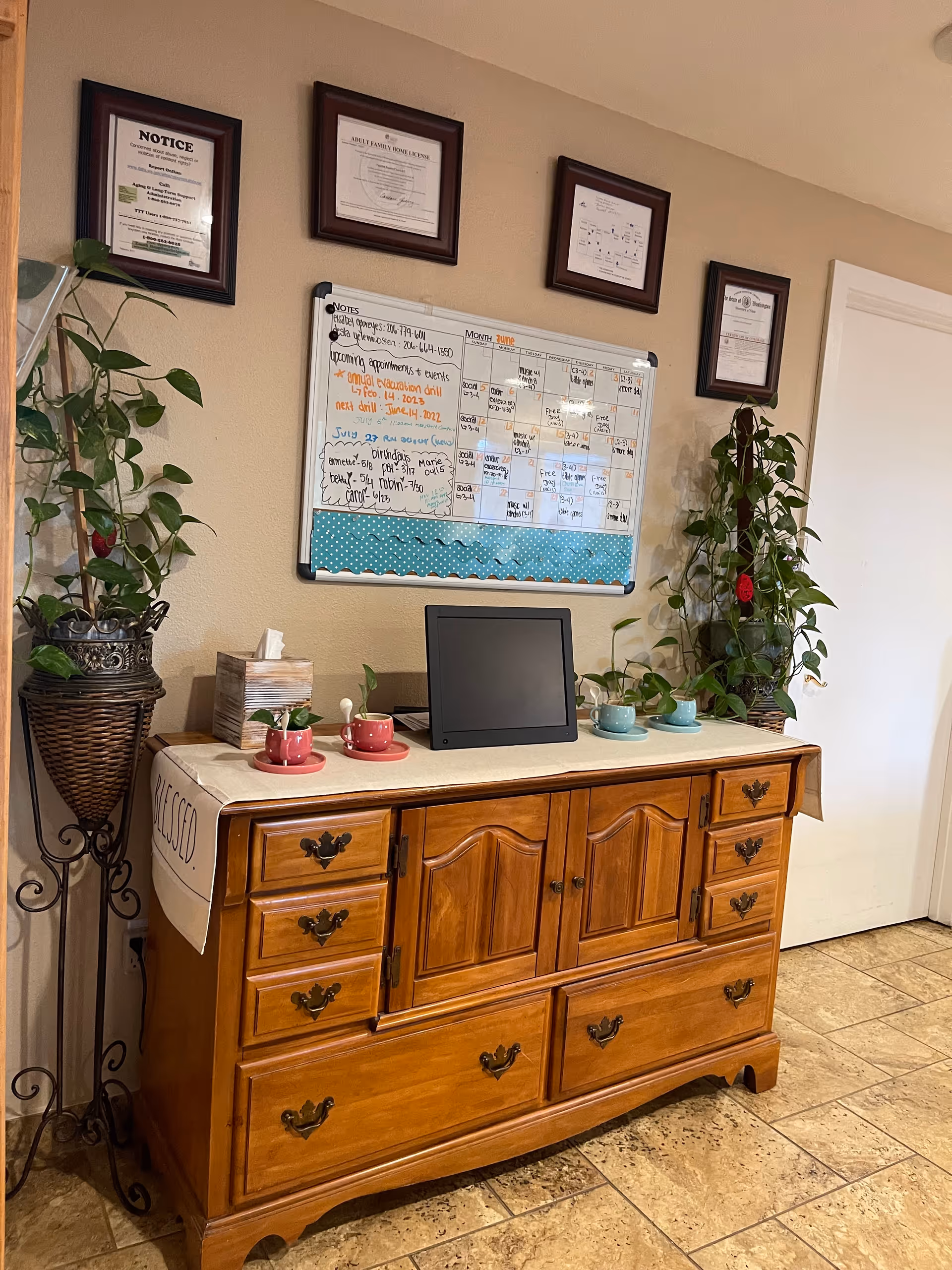 A wooden sideboard with multiple drawers and cabinets stands against a beige wall. On top of the sideboard are two sets of small cups and saucers in red and blue, a tissue box, a small plant, and a digital photo frame or tablet. Above the sideboard is a whiteboard calendar with notes and appointments written on it. Four framed certificates or notices hang on the wall above the whiteboard. There are two tall potted plants on either side of the sideboard. The floor is tiled in a light brown color.