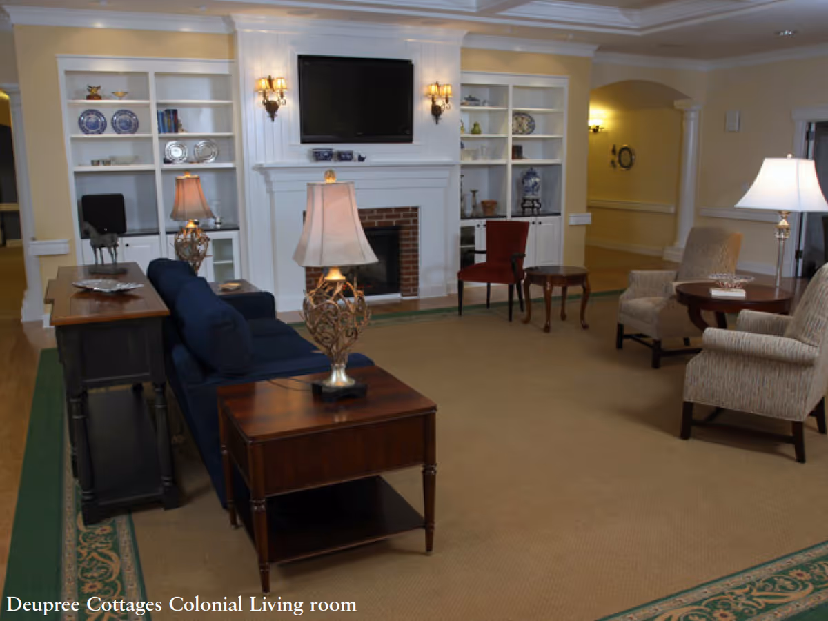 A cozy living room with a blue sofa, two beige armchairs, a red chair, wooden side tables with lamps, built-in white shelves with decorative items, a fireplace with a mounted TV above it, and warm lighting.