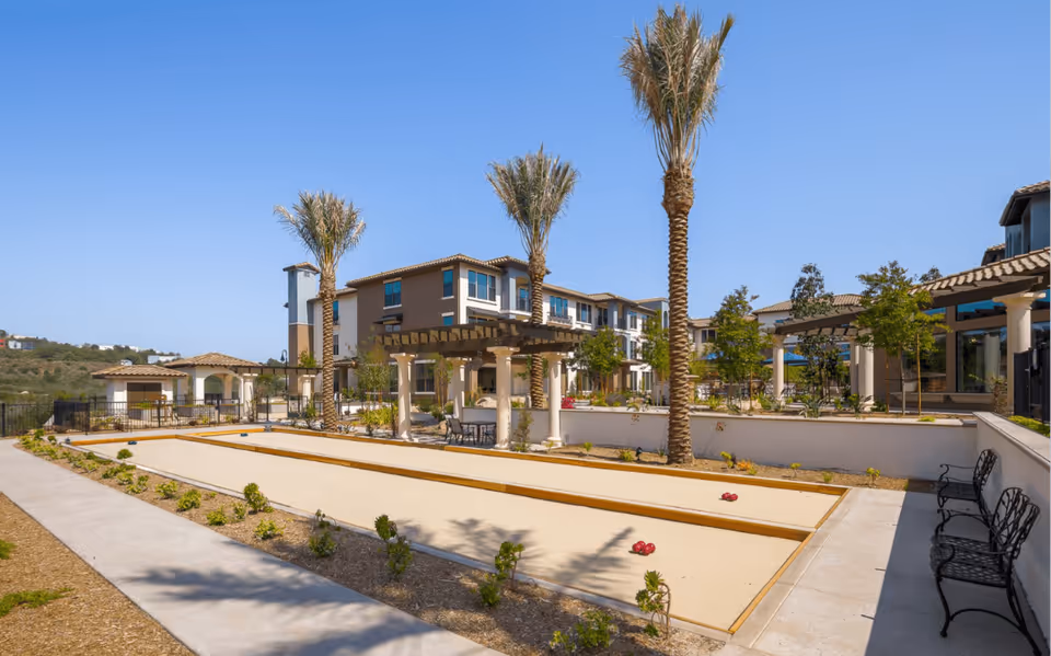 Outdoor recreational area at Santianna Oakmont Signature Senior Living featuring bocce ball courts, palm trees, benches, and pergolas with a multi-story residential building in the background under a clear blue sky.