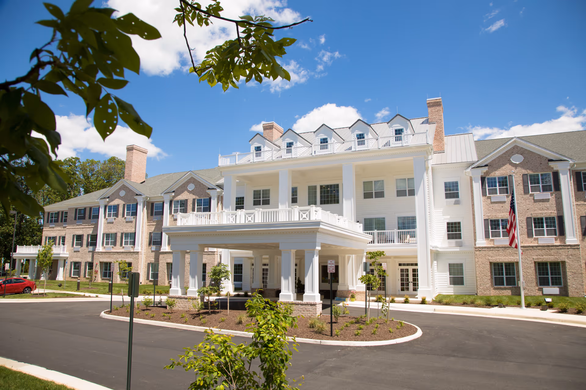 Front exterior view of a large, elegant senior living facility building with white columns, multiple windows, and a covered entrance. The building is surrounded by a paved driveway, landscaped greenery, and trees under a bright blue sky with some clouds.