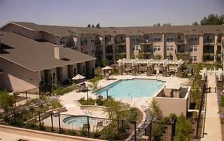 View of an outdoor courtyard area in a senior living facility featuring a large swimming pool surrounded by lounge chairs and umbrellas, a smaller hot tub, landscaped greenery, and multi-story residential buildings in the background under a clear sky.
