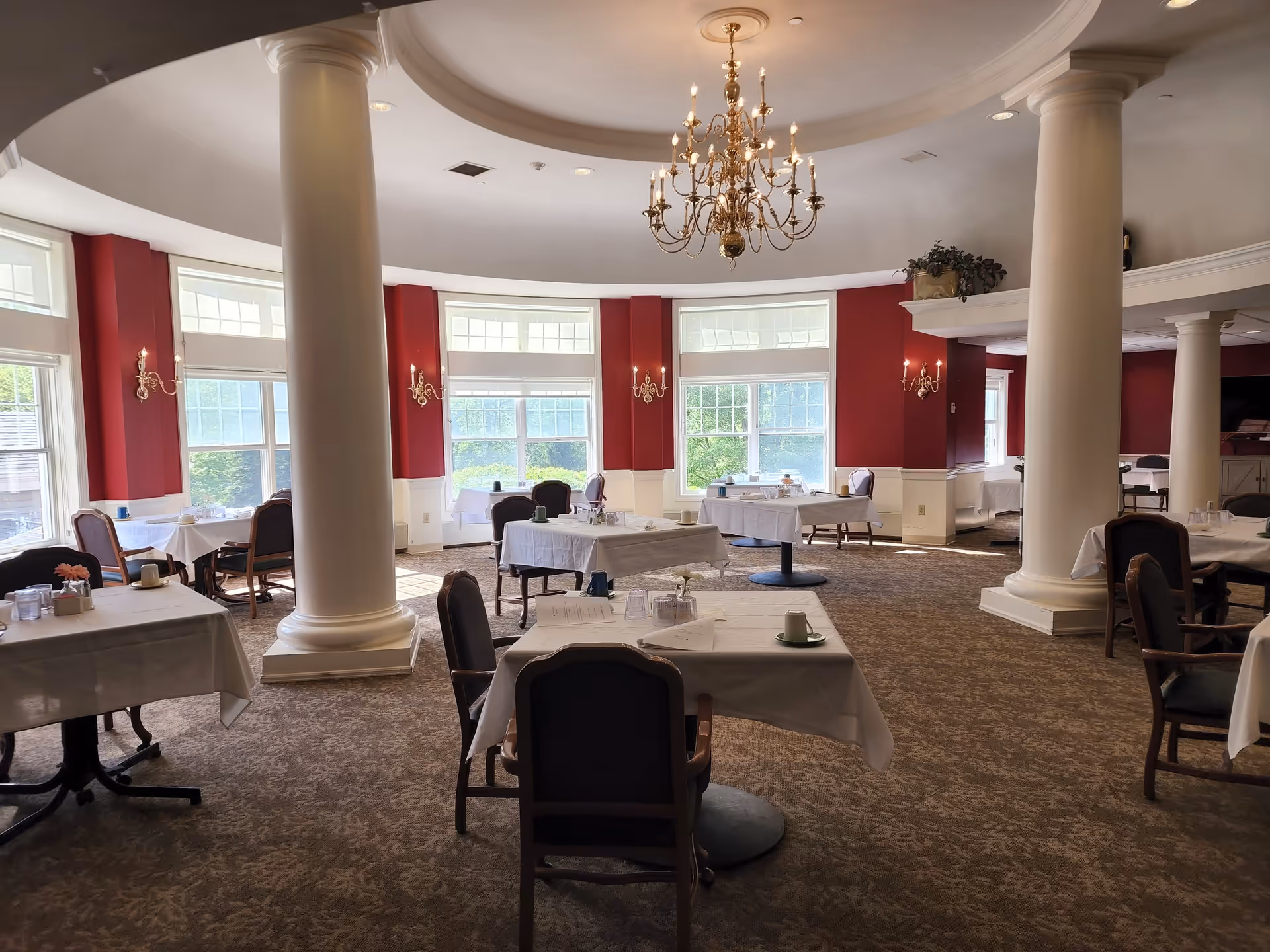 Dining room with white-tableclothed tables, upholstered chairs, large columns, and a central chandelier.