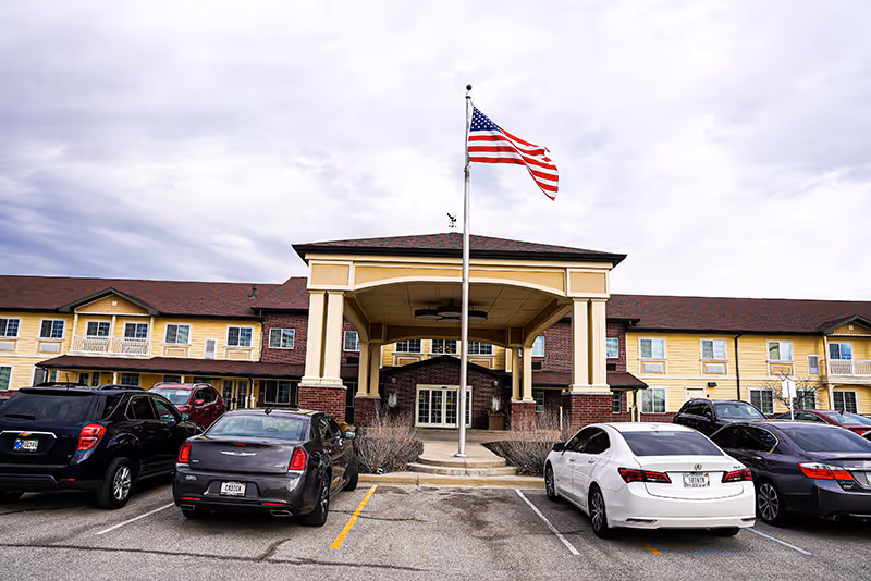 Front entrance of Meadow Brook Senior Living with an American flag on a pole and several cars parked in the lot.