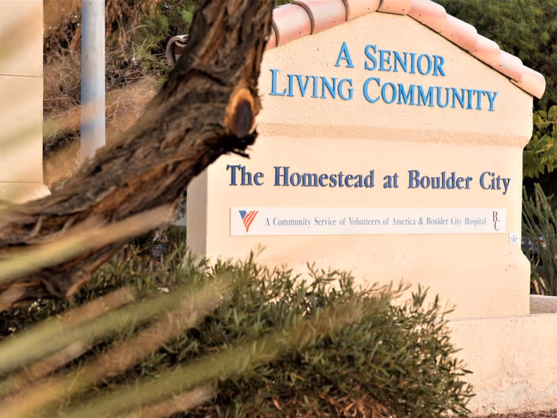 Stone entrance sign reading "A Senior Living Community - The Homestead at Boulder City" with landscaping and a tree trunk in the foreground.