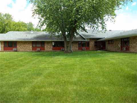 Single-story brick senior living facility with green lawn and a large tree in front.