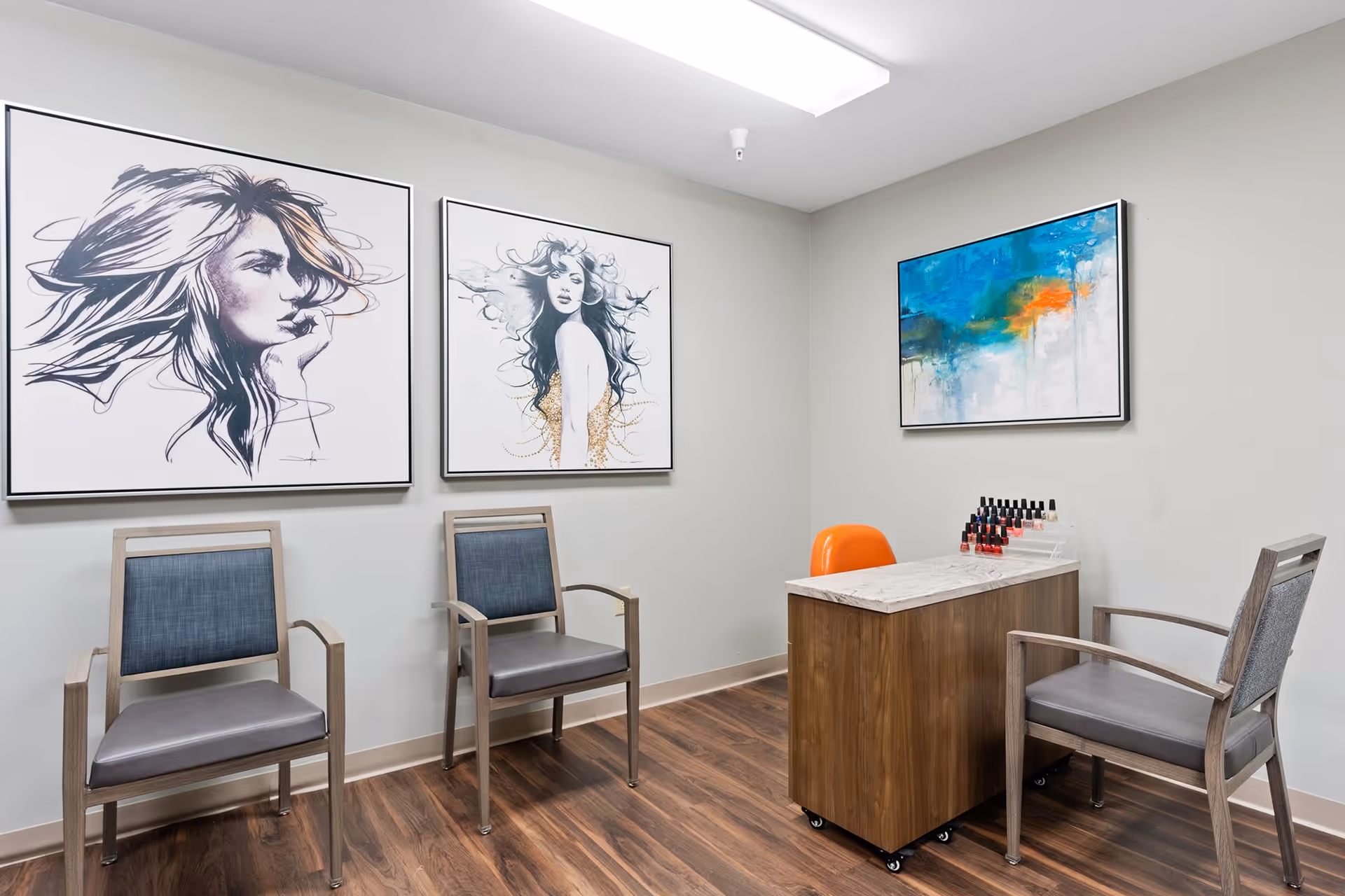 A small room with three chairs and a wooden table with a marble top. On the table are several bottles of nail polish. The walls are decorated with three framed artworks, two featuring stylized portraits of women and one abstract painting with blue, green, and orange colors. The floor is wood, and the room is lit by a ceiling light.