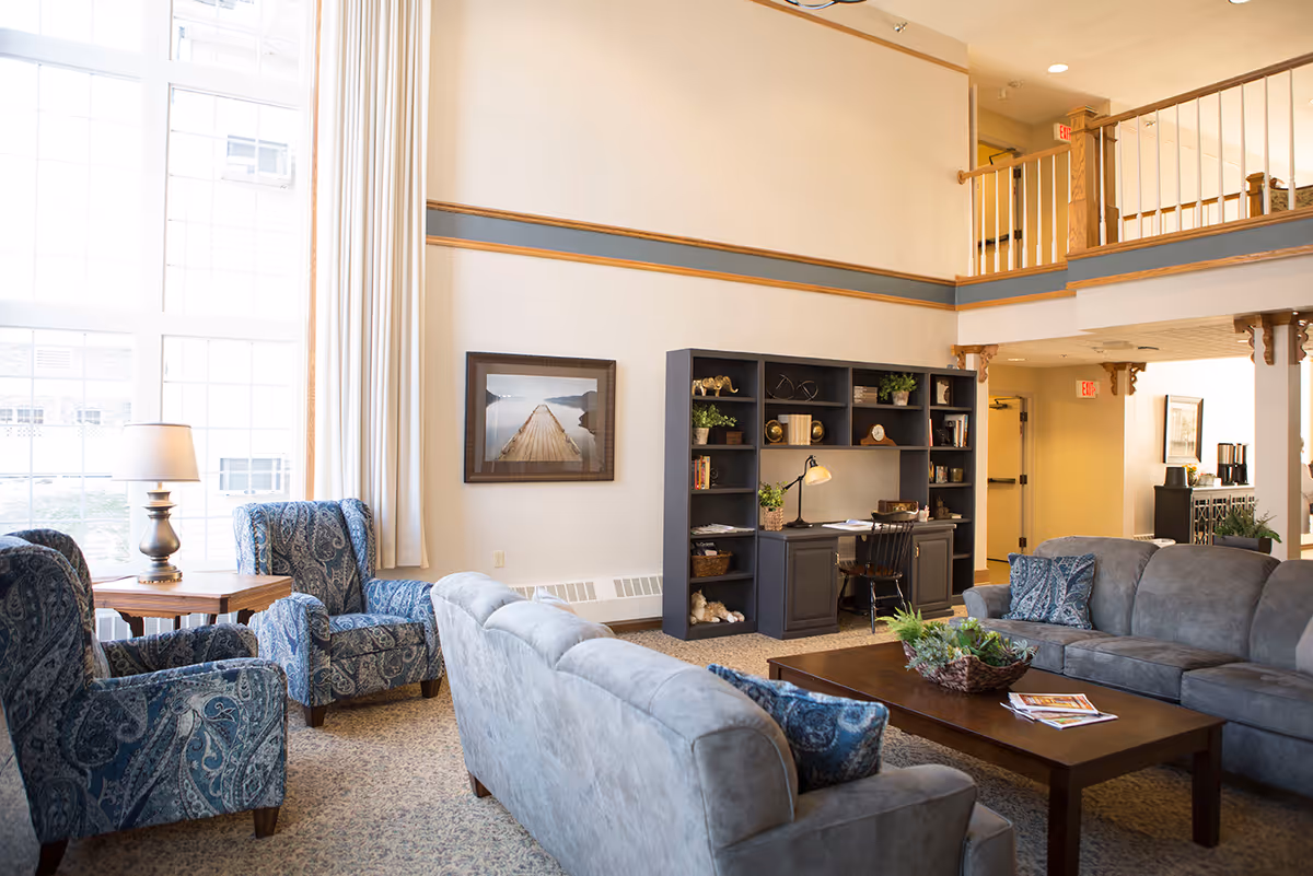 A bright and spacious living room area with large windows letting in natural light. The room features two blue patterned armchairs, two gray sofas with blue patterned pillows, a wooden coffee table with a plant centerpiece and magazines, and a dark wood desk with shelves filled with books and decorative items. The walls are light-colored with a framed picture of a wooden pier over water. There is a balcony railing visible on the upper level and an exit sign near a doorway in the background.