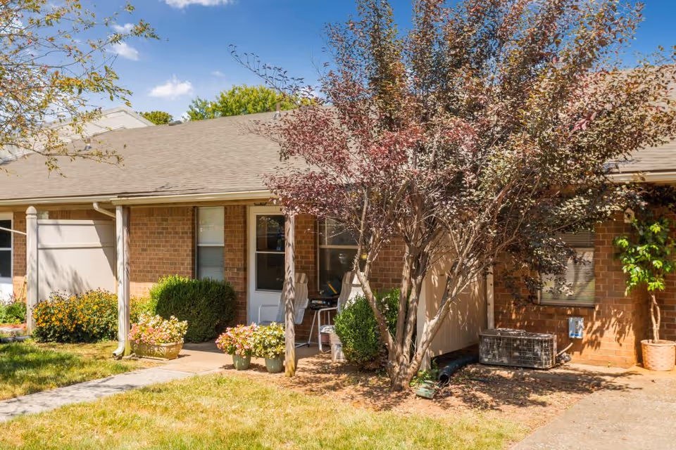 Exterior view of a single-story brick building with a small covered porch area. There are potted plants and bushes near the entrance, a tree with reddish leaves providing shade, and a clear blue sky above.