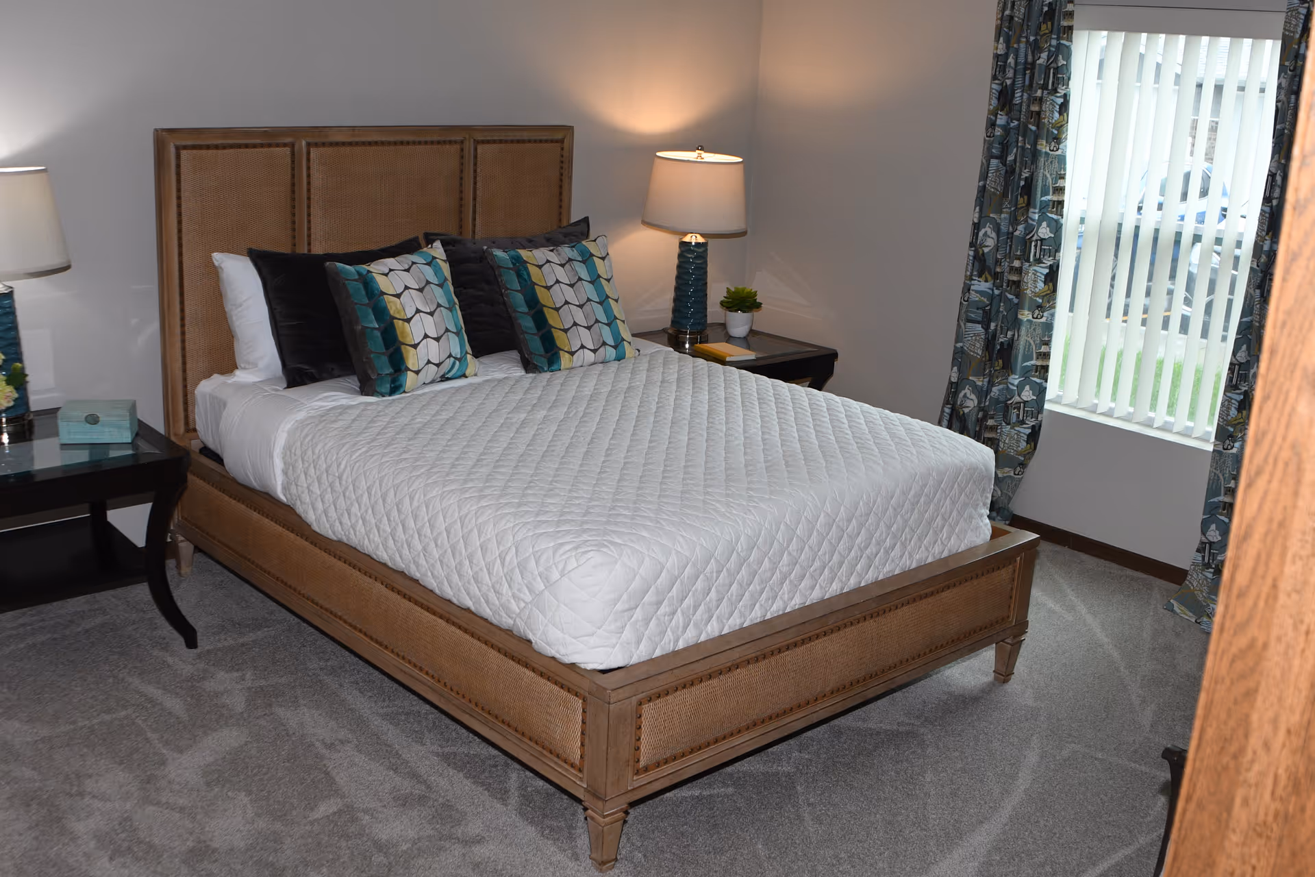 A furnished bedroom with a wooden bed, white quilted bedding, patterned pillows, nightstands with lamps, and a window with vertical blinds.
