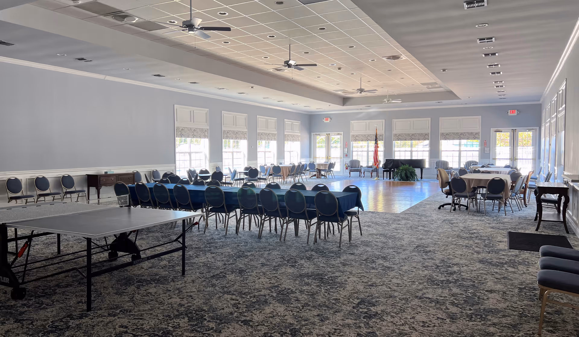 A large, well-lit multipurpose room with several tables and chairs arranged around the space. There is a ping pong table in the foreground, a piano near the far wall, and an American flag next to it. The room has multiple windows letting in natural light, ceiling fans, and a patterned carpeted floor.