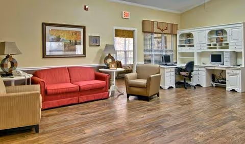 A cozy common area with a red sofa, two beige armchairs, a white desk with a computer and office chair, two table lamps, framed artwork on the wall, and large windows with blinds. The room has wood flooring and light-colored walls.