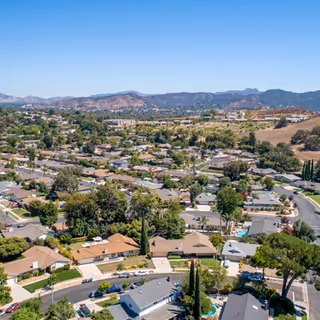 Aerial view of a suburban neighborhood with single-family homes, yards, pools and rolling hills in the background under a clear sky.