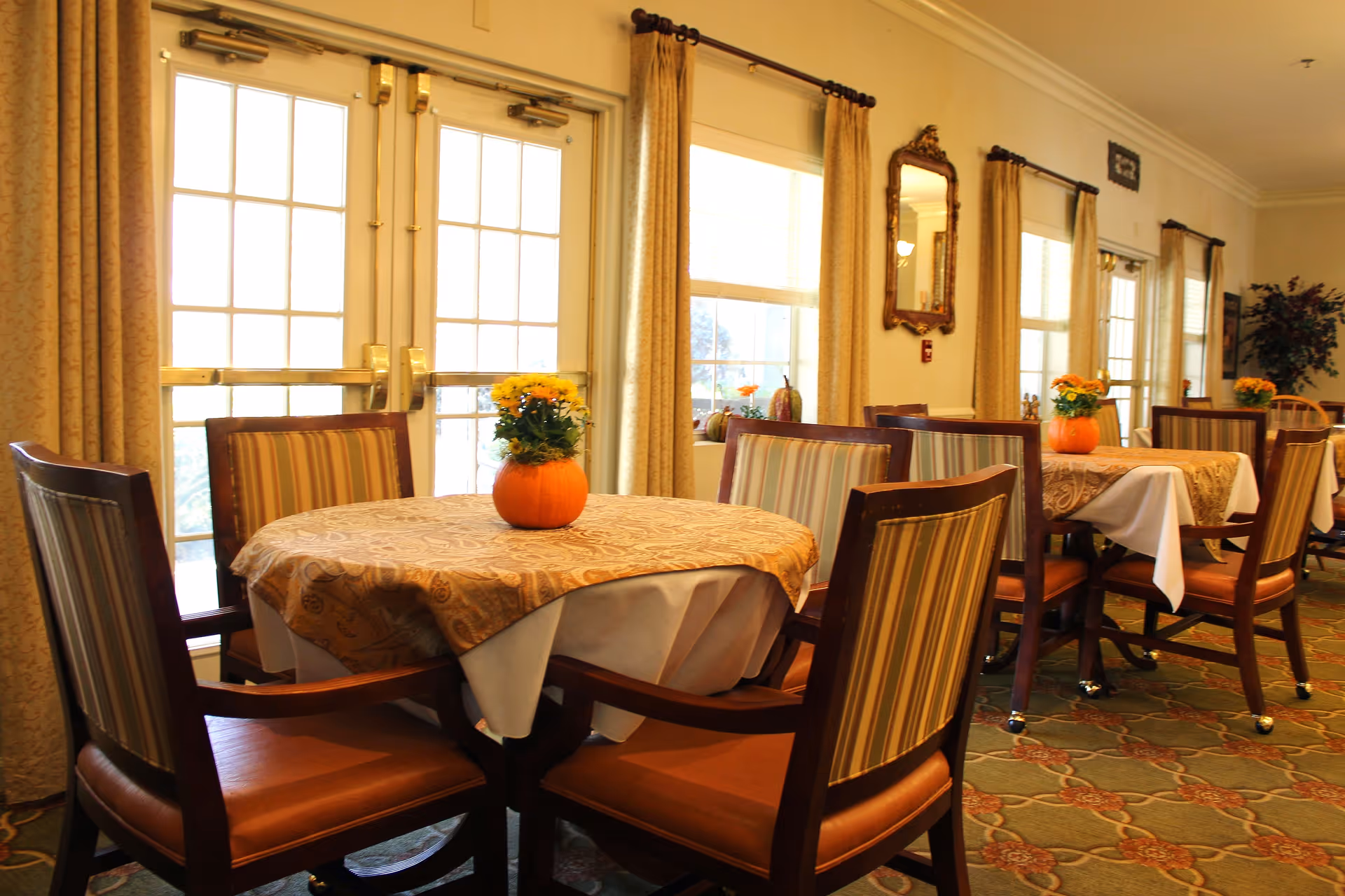 A bright dining room with round tables covered in patterned tablecloths and white underlays, each table decorated with a small pumpkin vase holding yellow flowers. The room features large windows with beige curtains, wooden chairs with striped upholstery, and a patterned carpet. A mirror and some wall decorations are visible on the cream-colored walls.