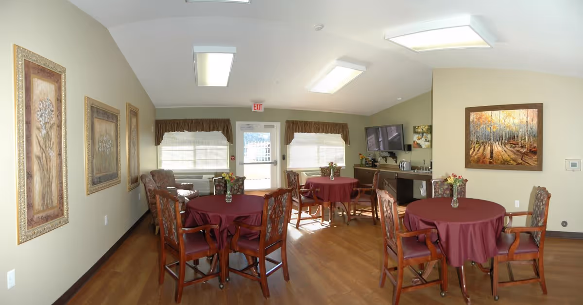 A well-lit common area with round tables covered in burgundy tablecloths, each with a vase of flowers. Wooden chairs with patterned cushions surround the tables. The room has light-colored walls adorned with framed artwork and large windows with valances. A television is mounted on the wall above a counter with a sink and coffee maker. The floor is wooden, and there is an exit door at the back of the room.