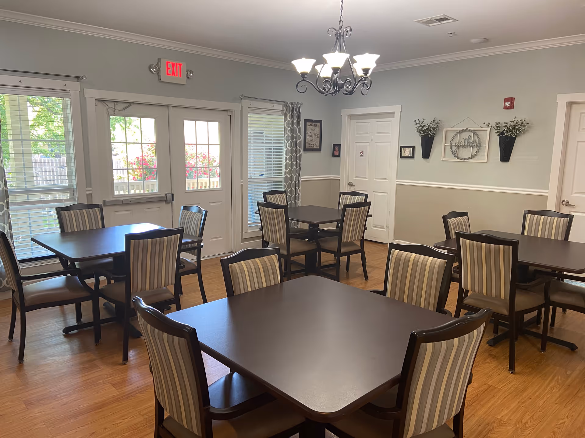 A dining room with several dark wood tables and striped cushioned chairs. There are large windows and glass doors letting in natural light, with an exit sign above the doors. The walls are painted light gray with white trim, decorated with framed pictures and wall-mounted planters with greenery. A chandelier with multiple lights hangs from the ceiling.
