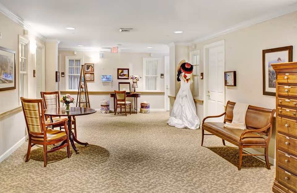 A well-lit hallway in a senior living facility featuring a small round table with two wooden chairs on the left, a wooden bench with a throw blanket on the right, and a mannequin dressed in a white gown with hats hanging on it near the back wall. The walls are decorated with framed pictures, and there is a wooden chest of drawers on the right side.