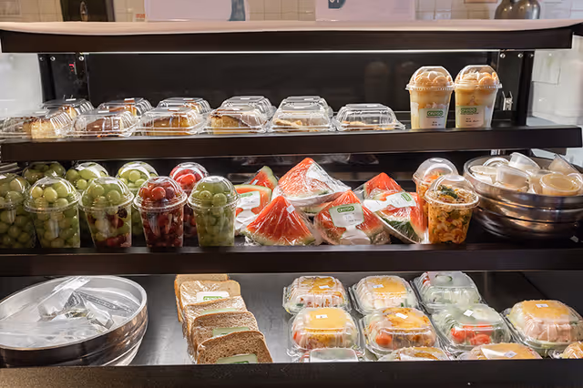 A refrigerated display case with three shelves containing various pre-packaged food items including sandwiches, fruit cups with grapes and cherries, slices of watermelon, salads, and desserts in clear plastic containers.