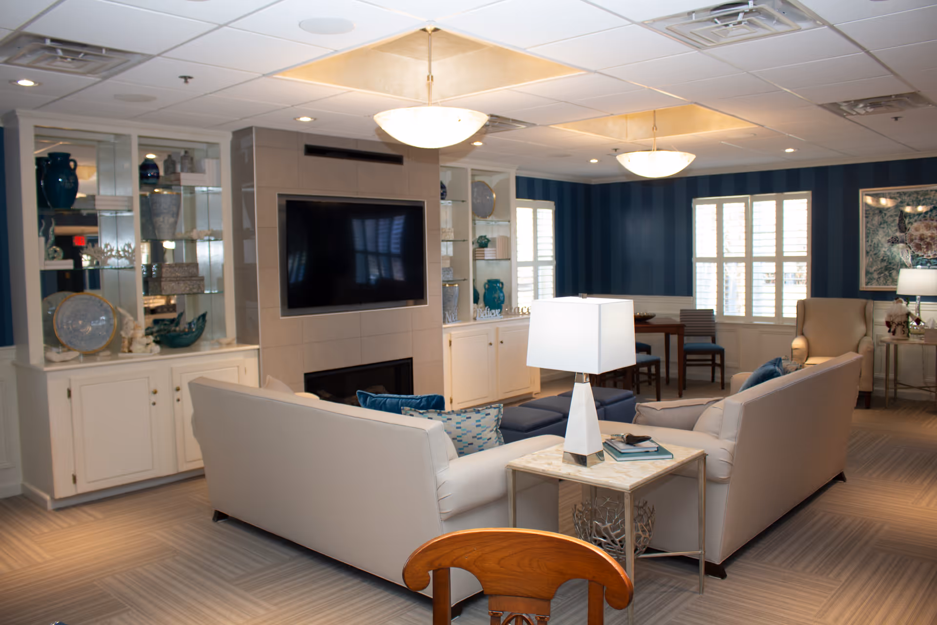 A furnished senior living lounge with two sofas facing a wall-mounted TV above a fireplace, built-in shelving, lamps, and a small dining area by the windows.