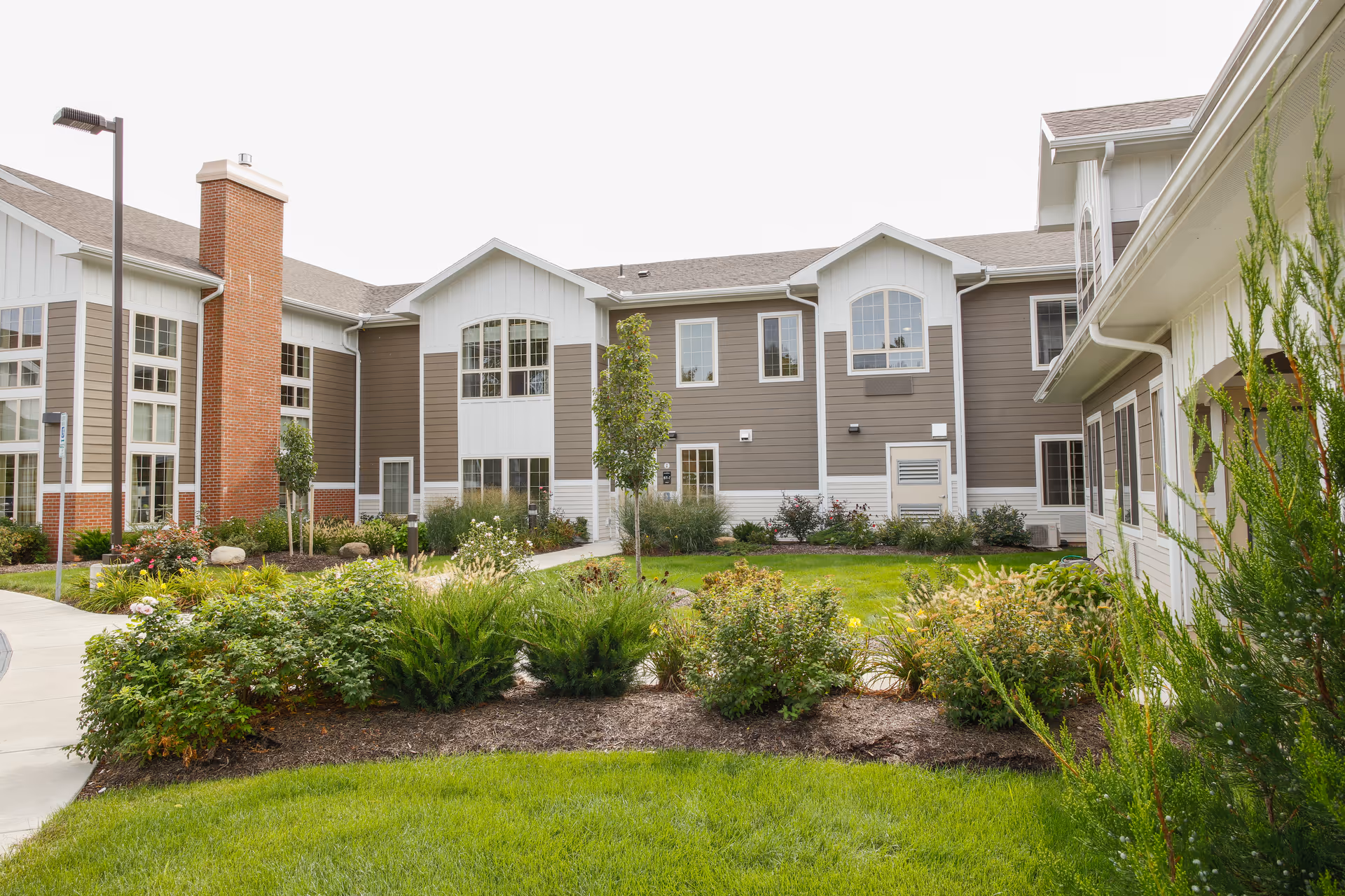 Exterior view of a senior living facility building with beige and white siding, large windows, a brick chimney, and a landscaped garden with green grass, bushes, and small trees in the foreground.