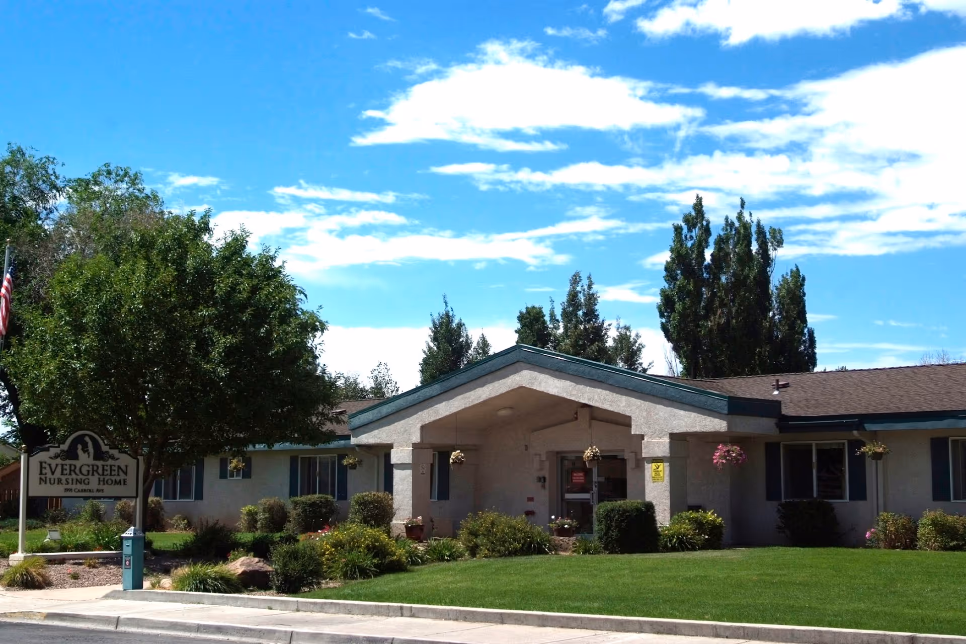 Exterior view of Evergreen Nursing Home building with a well-maintained lawn, trees, and shrubs under a partly cloudy blue sky.