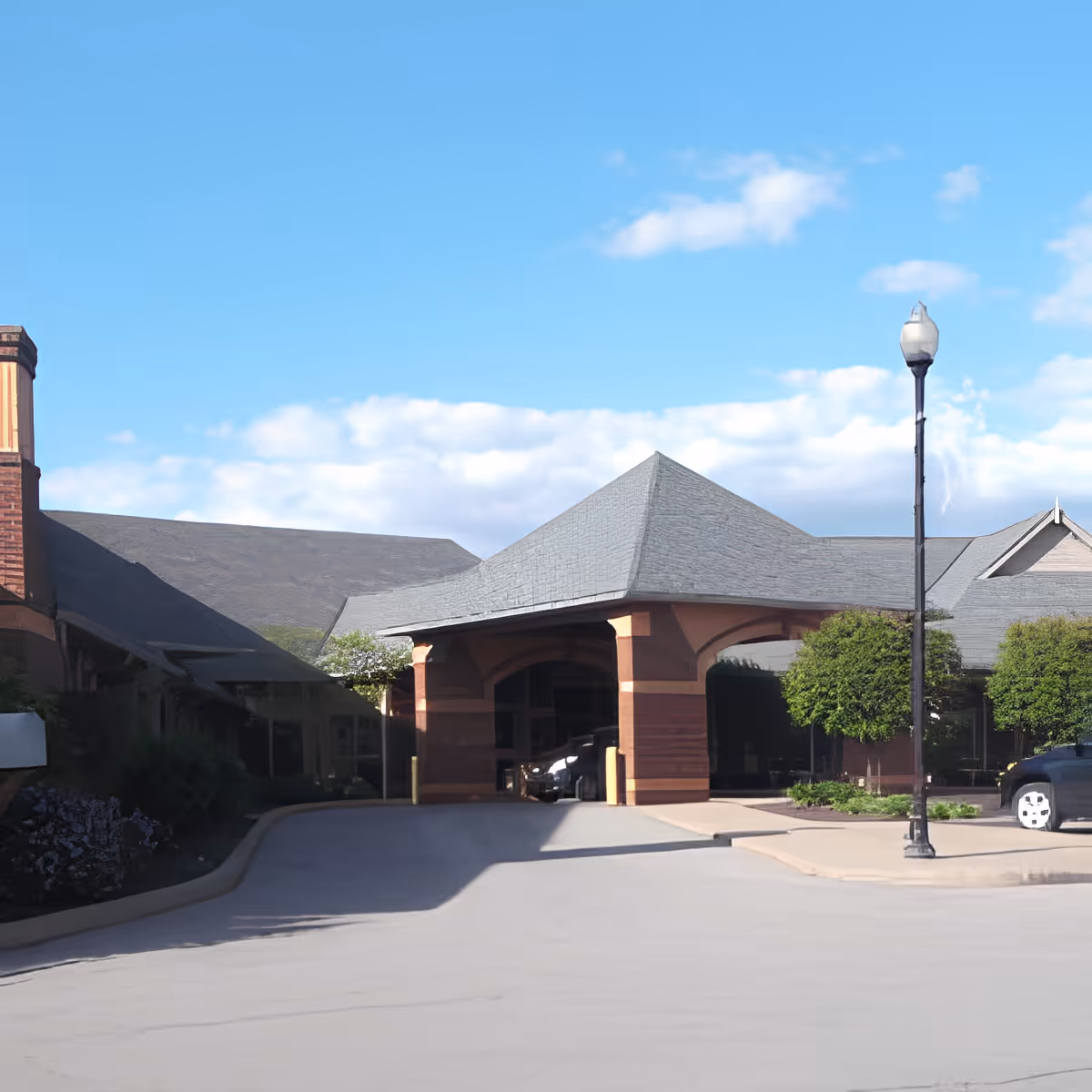 Covered entrance/porte-cochère of a brick-clad senior living facility with landscaping and a lamp post under a blue sky.
