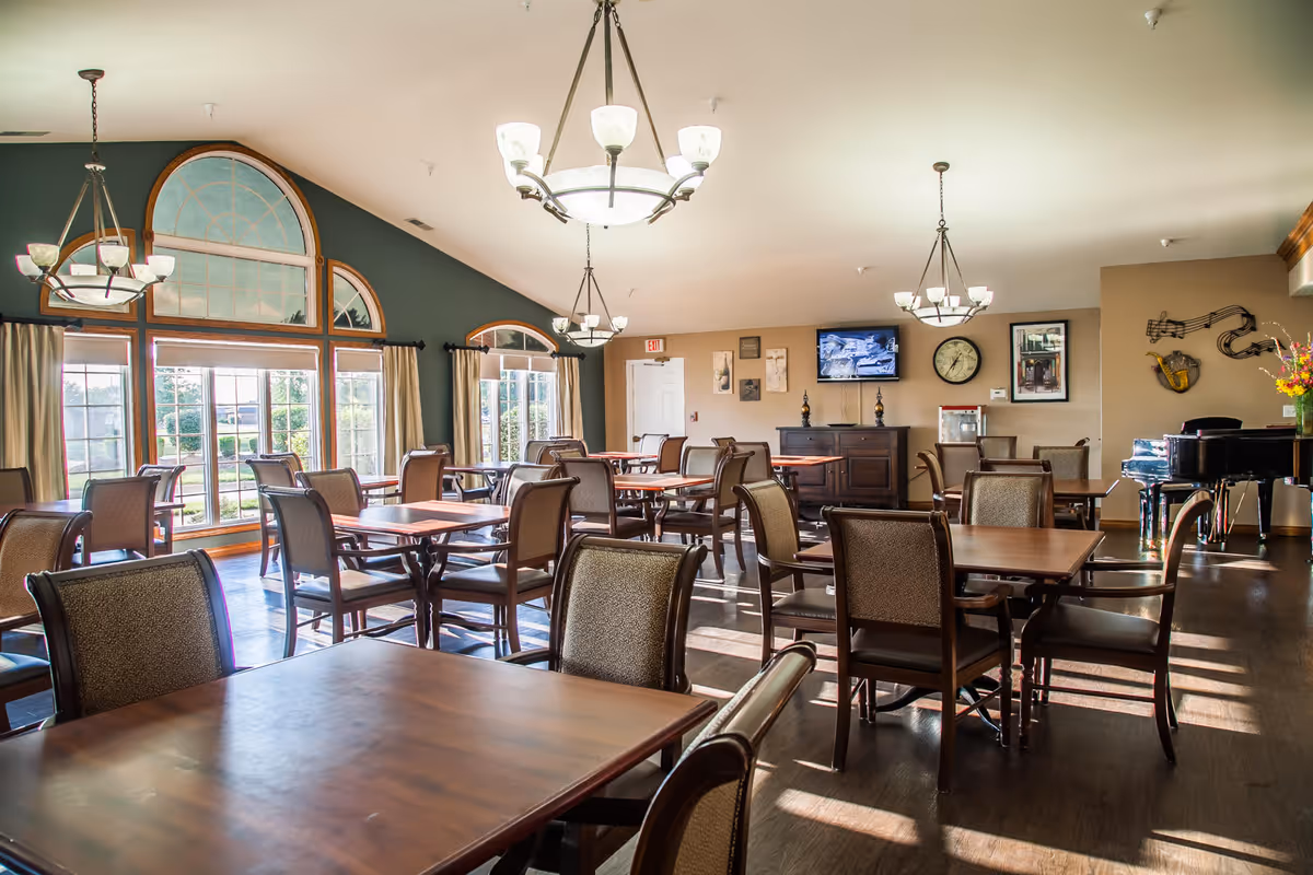 A bright and spacious dining room with multiple wooden tables and cushioned chairs arranged neatly. Large windows with arched tops allow natural light to fill the room. The walls are decorated with framed pictures, a clock, and musical instrument wall art. A black grand piano is placed in the corner, and several hanging light fixtures illuminate the space.