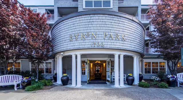 Front exterior view of Byron Park facility with a curved entrance supported by white columns, flanked by purple-leafed trees and benches on either side.