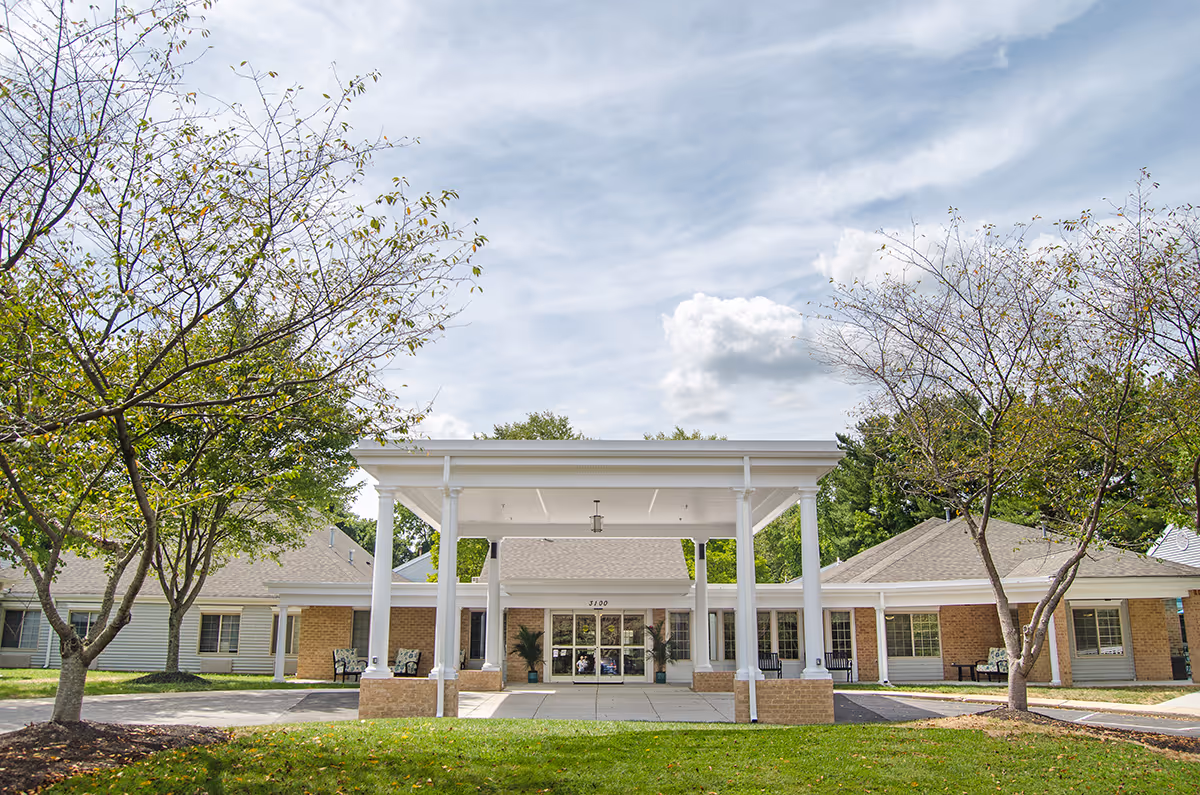 Front exterior view of Lighthouse Senior Living at Ellicott City showing a single-story building with a covered entrance supported by white columns, surrounded by trees and a green lawn under a partly cloudy sky.