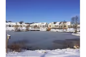 Snow-covered pond in the foreground with a row of light-colored residential buildings and a clear blue sky.