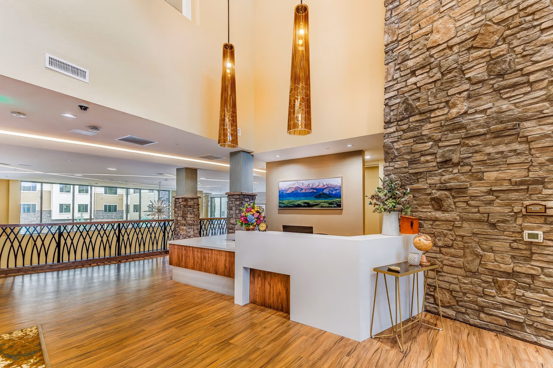 A modern reception area with a white and wood front desk, two tall pendant lights hanging from a high ceiling, a stone accent wall, and a painting of mountains on the wall behind the desk. There is a small table with decorative items including a globe and flowers next to the desk. Large windows and a railing are visible in the background.