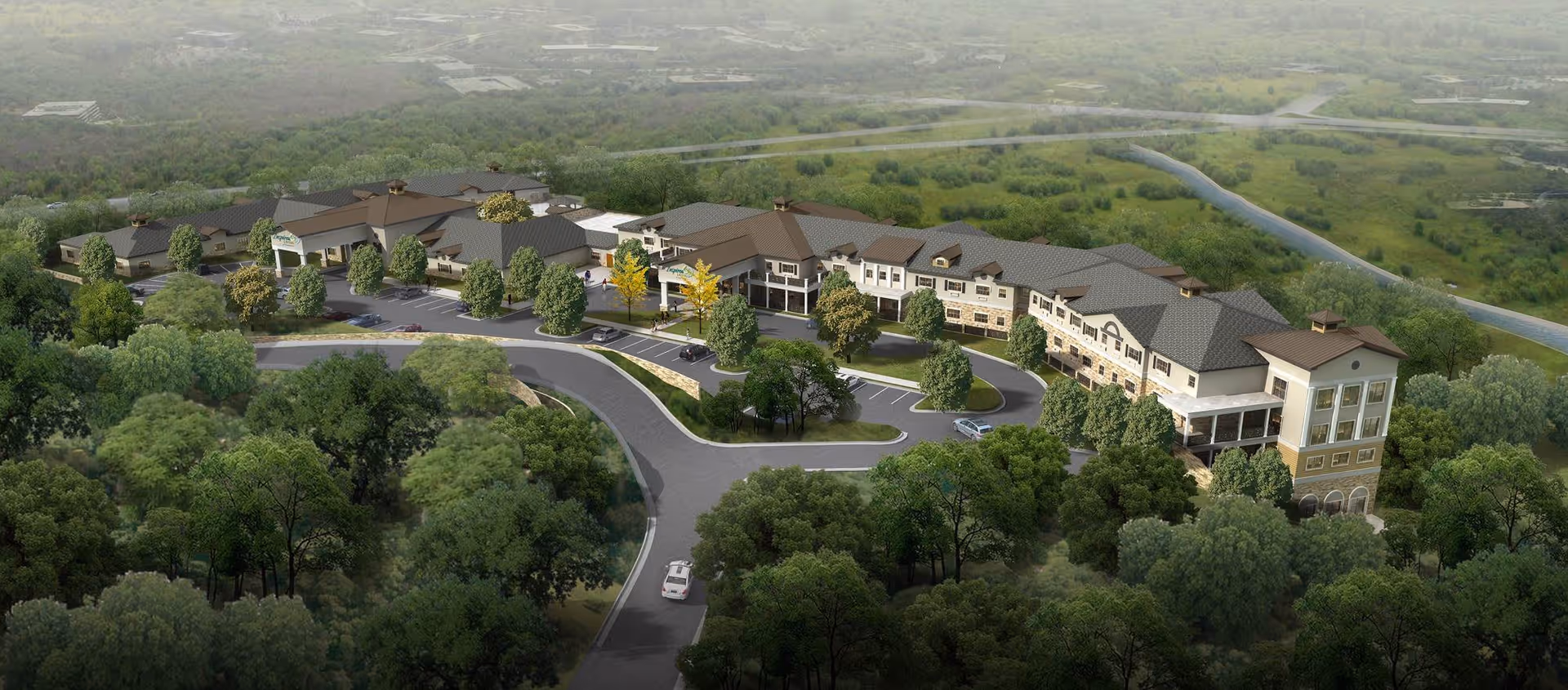 Aerial view of a large senior living facility surrounded by dense green trees and natural landscape. The building has multiple sections with a mix of architectural styles, including pitched roofs and stone accents. There is a curved driveway and parking area in front of the building, with several cars parked. The facility is set in a lush, wooded area with rolling hills in the background.