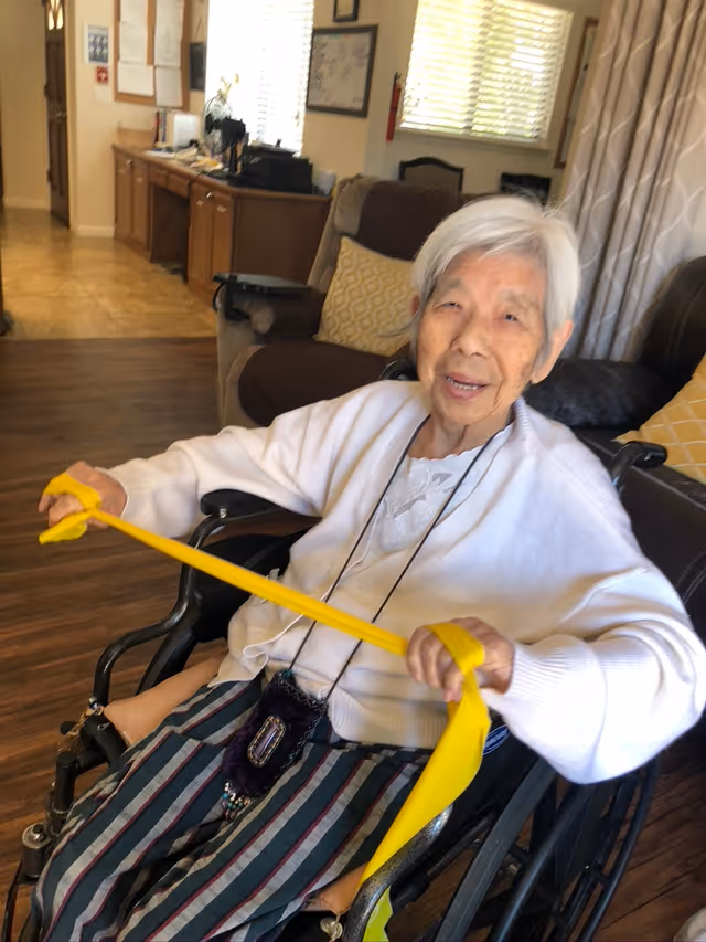 An elderly woman with white hair sitting in a wheelchair inside a living room area, smiling and holding a yellow exercise resistance band stretched between her hands. Behind her are a brown couch with yellow patterned pillows, a window with blinds, and a wooden cabinet with various items on top.