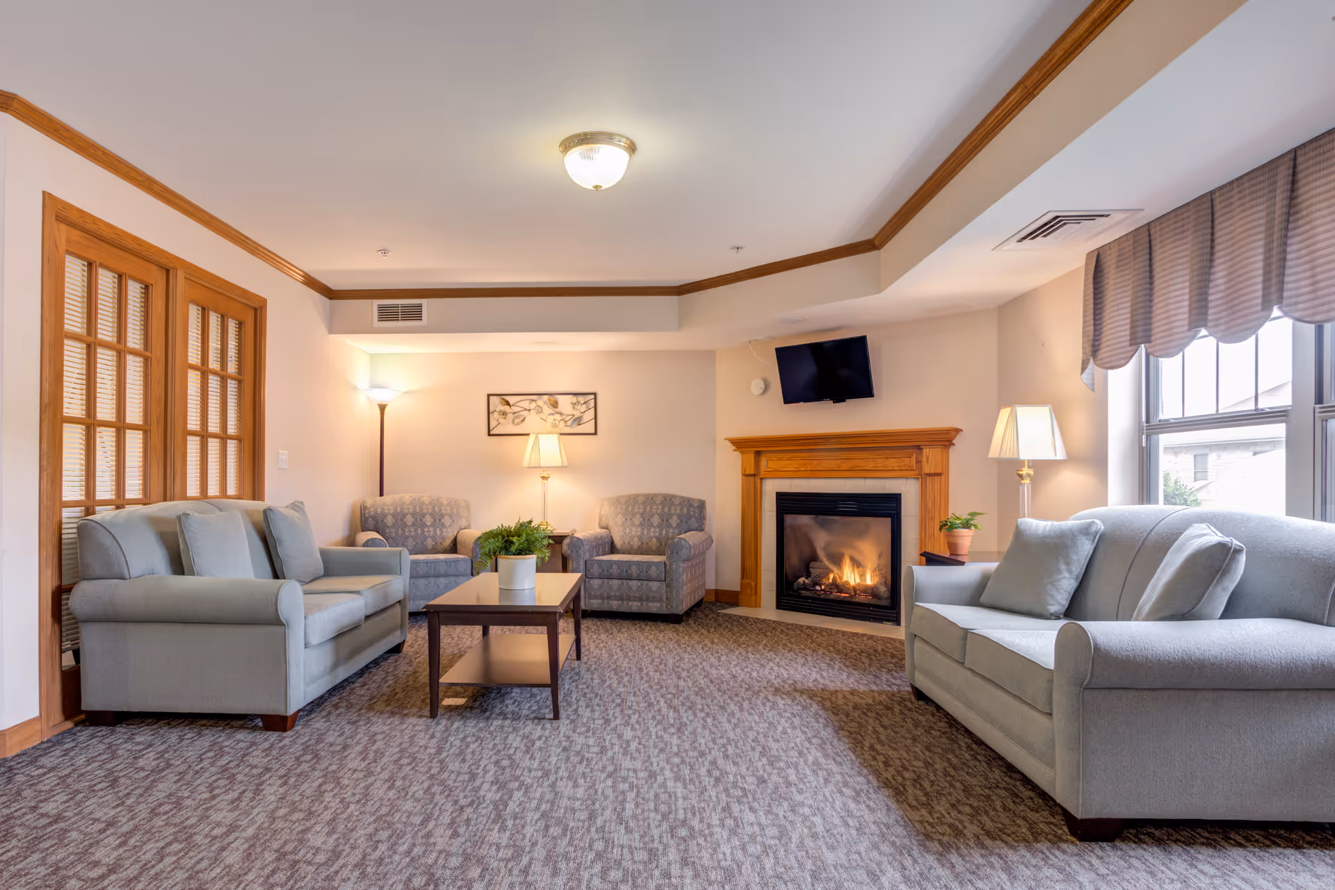 A cozy living room with two gray sofas and two patterned armchairs arranged around a wooden coffee table. There is a lit fireplace with a wooden mantel, a wall-mounted TV above it, and two lamps providing warm lighting. A large window with striped valances lets in natural light.