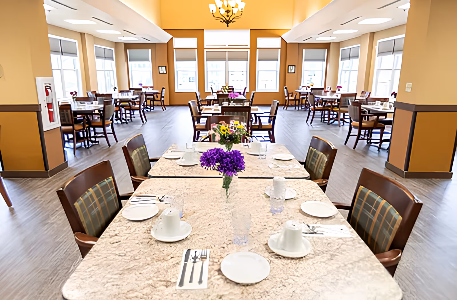 A spacious dining room with multiple tables and chairs arranged neatly. Each table is set with white plates, cups, and silverware, and decorated with small flower arrangements. Large windows allow natural light to fill the room, and the walls are painted in warm beige tones. The floor is covered with wood-style flooring, and a chandelier hangs from the ceiling.
