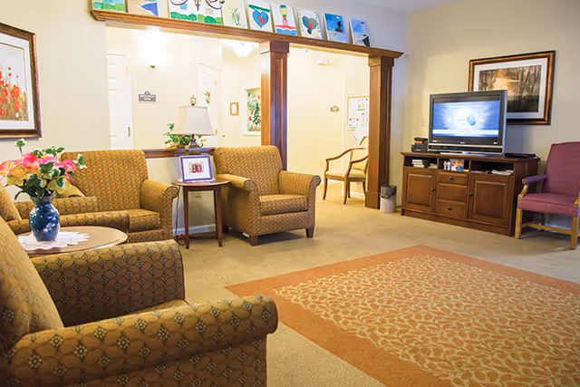 A cozy living room area with patterned armchairs and a sofa arranged around a large area rug. A wooden TV stand holds a flat-screen television, and there is a framed picture on the wall. A small side table with a lamp and a framed photo is placed between two armchairs. The room has soft lighting and a warm, inviting atmosphere with artwork displayed on a ledge above a wooden beam.