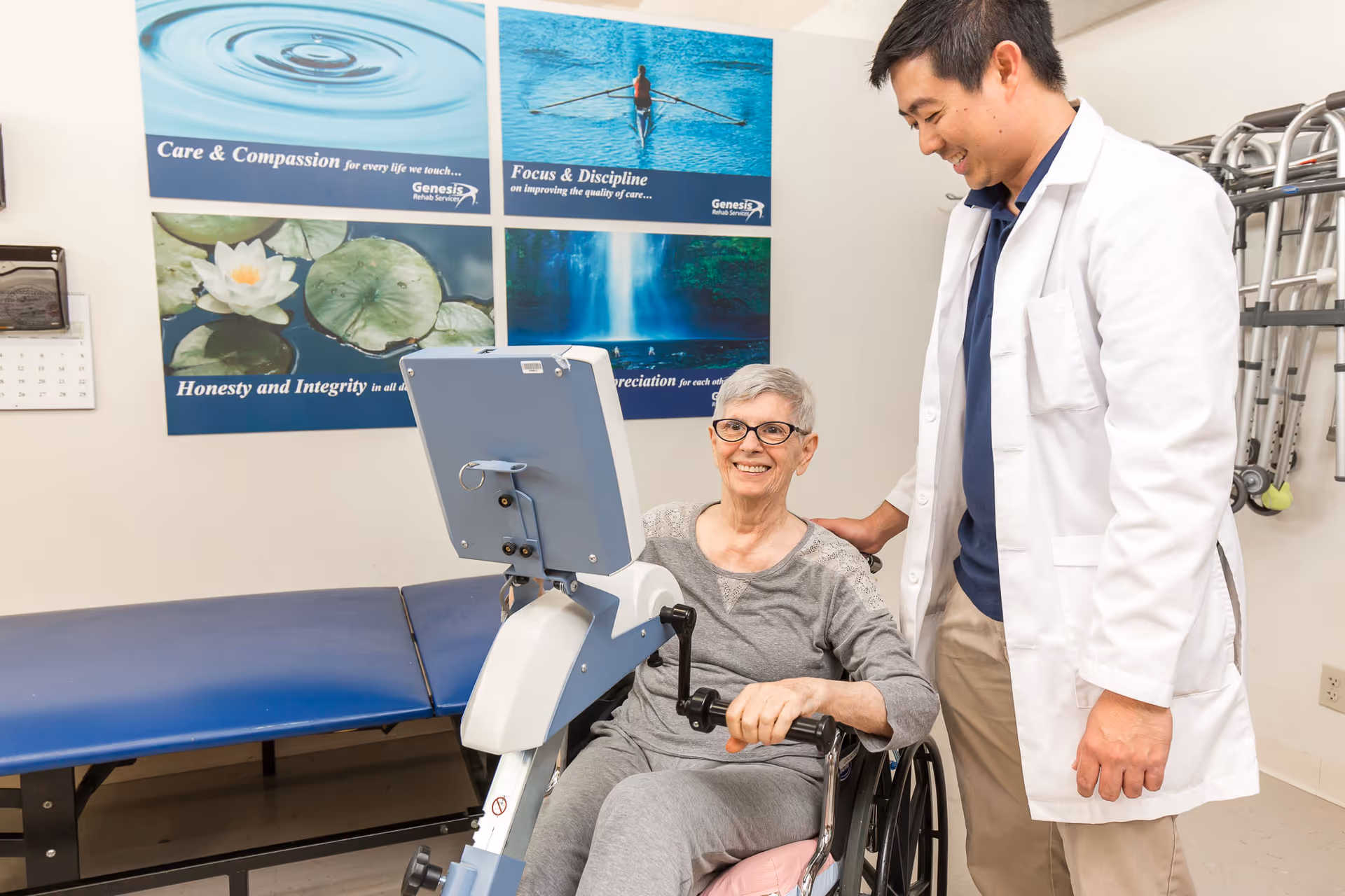 An elderly woman in a wheelchair using a hand cycle exercise machine while a male healthcare professional in a white coat stands beside her, smiling. The room has motivational posters on the wall and exercise equipment in the background.
