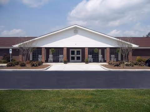 Front exterior view of a single-story brick building with a white gabled roof and a covered entrance. The entrance has double glass doors flanked by windows and rocking chairs on the porch. There are small landscaped areas with shrubs and trees on either side of the walkway leading to the entrance.