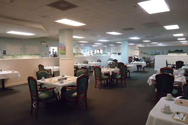 Large dining room with multiple round and rectangular tables set with white tablecloths and green-upholstered chairs in an assisted living facility.