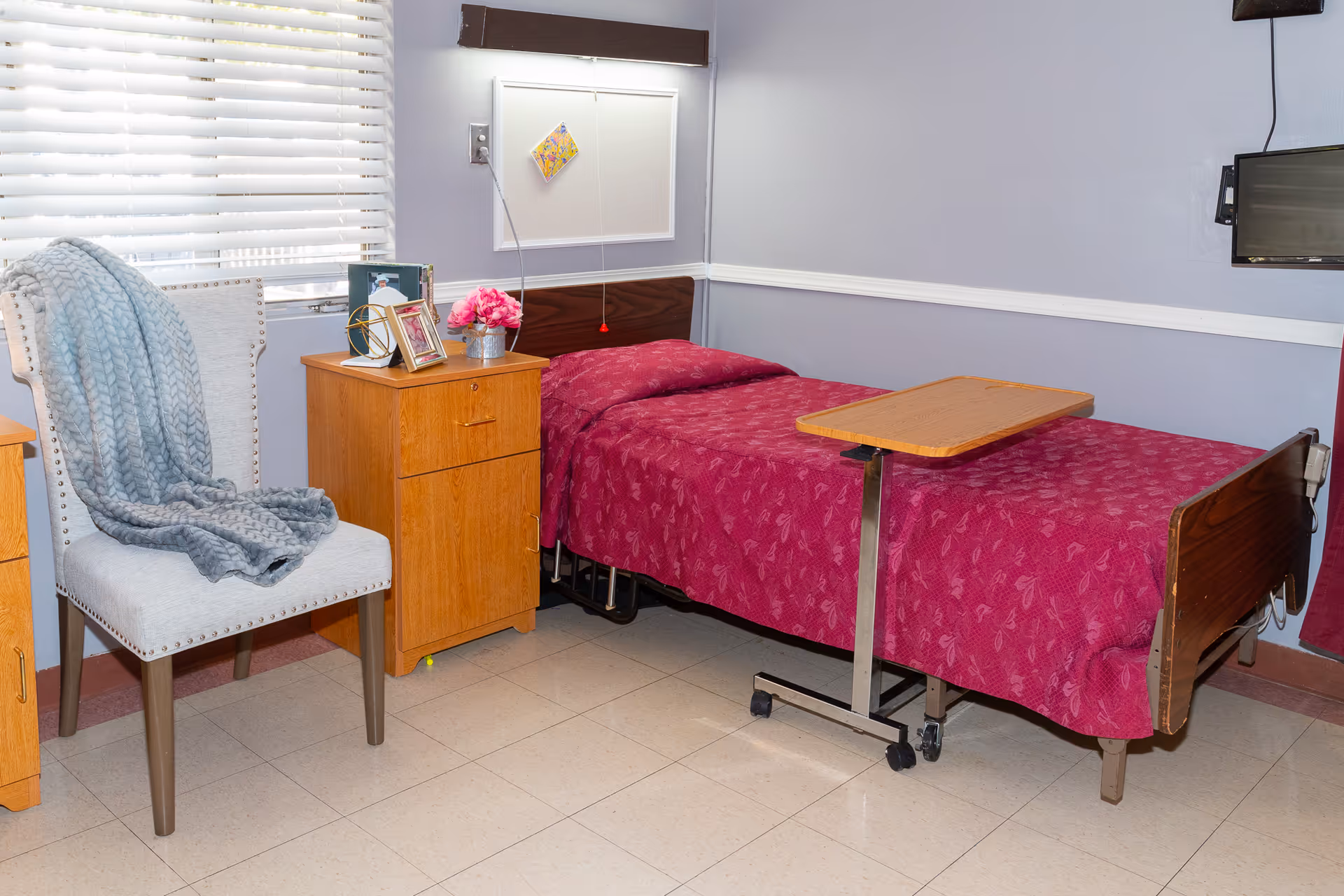 A small bedroom in a healthcare facility featuring a single bed with a red patterned blanket, a wooden bedside cabinet with framed photos and a flower vase, a light gray upholstered chair with a blue knitted blanket draped over it, a window with white blinds, and a mounted TV on the wall.