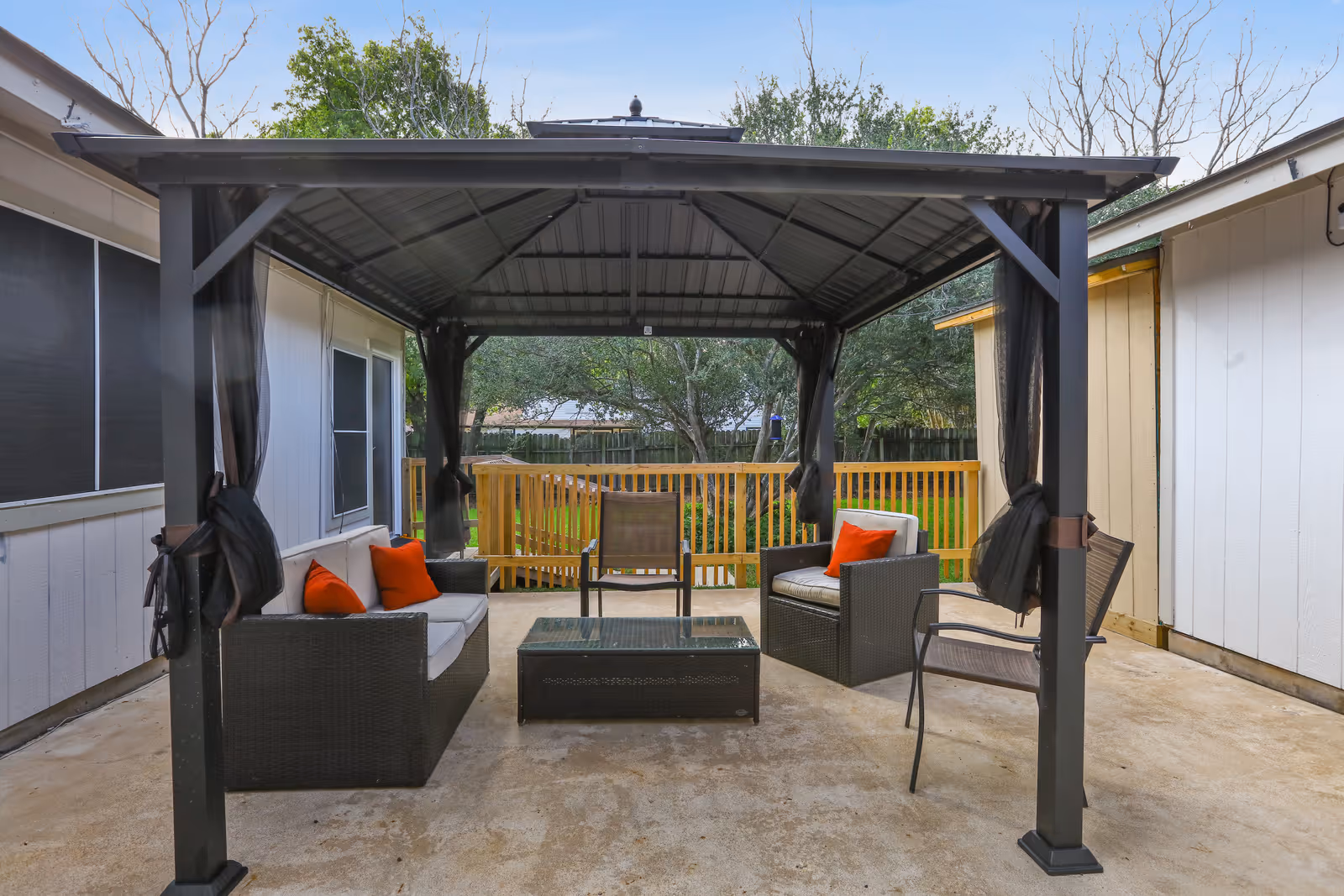 Outdoor covered patio area with a metal roof structure, featuring a seating arrangement including a sofa with orange cushions, two armchairs, a glass-top coffee table, and a single chair. The patio is surrounded by a wooden fence and trees in the background.
