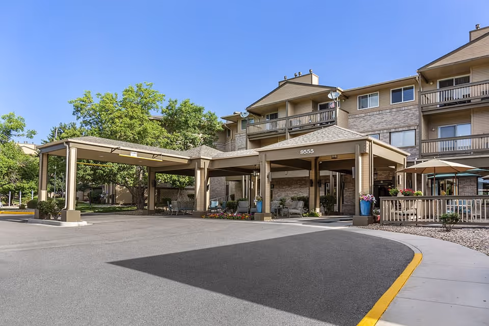 Covered porte-cochere and entrance of a multi-story senior living building with balconies, seating areas, and landscaped grounds under a clear blue sky.