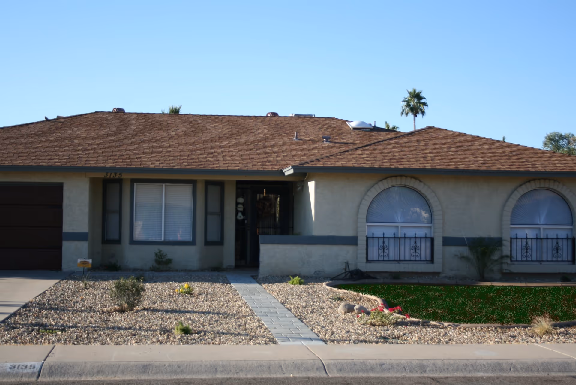 Single-story residential building with a brown shingle roof, beige exterior walls, and a front yard covered with gravel and some small plants. The building has a garage on the left, a central entrance door, and two large arched windows with decorative metal bars on the right. A paved walkway leads from the sidewalk to the front door. Palm trees are visible in the background under a clear blue sky.
