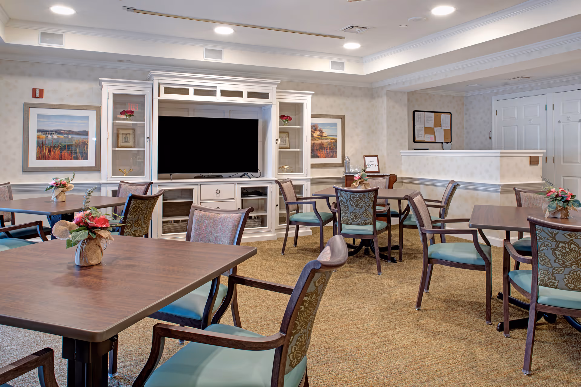 A well-lit common area in a senior living facility featuring several wooden tables with floral centerpieces and cushioned chairs arranged around them. At the back, there is a white entertainment center with a large flat-screen TV, glass cabinets with decorative items, and framed artwork on the walls. The room has beige carpeting and light-colored wallpaper, creating a cozy and inviting atmosphere.