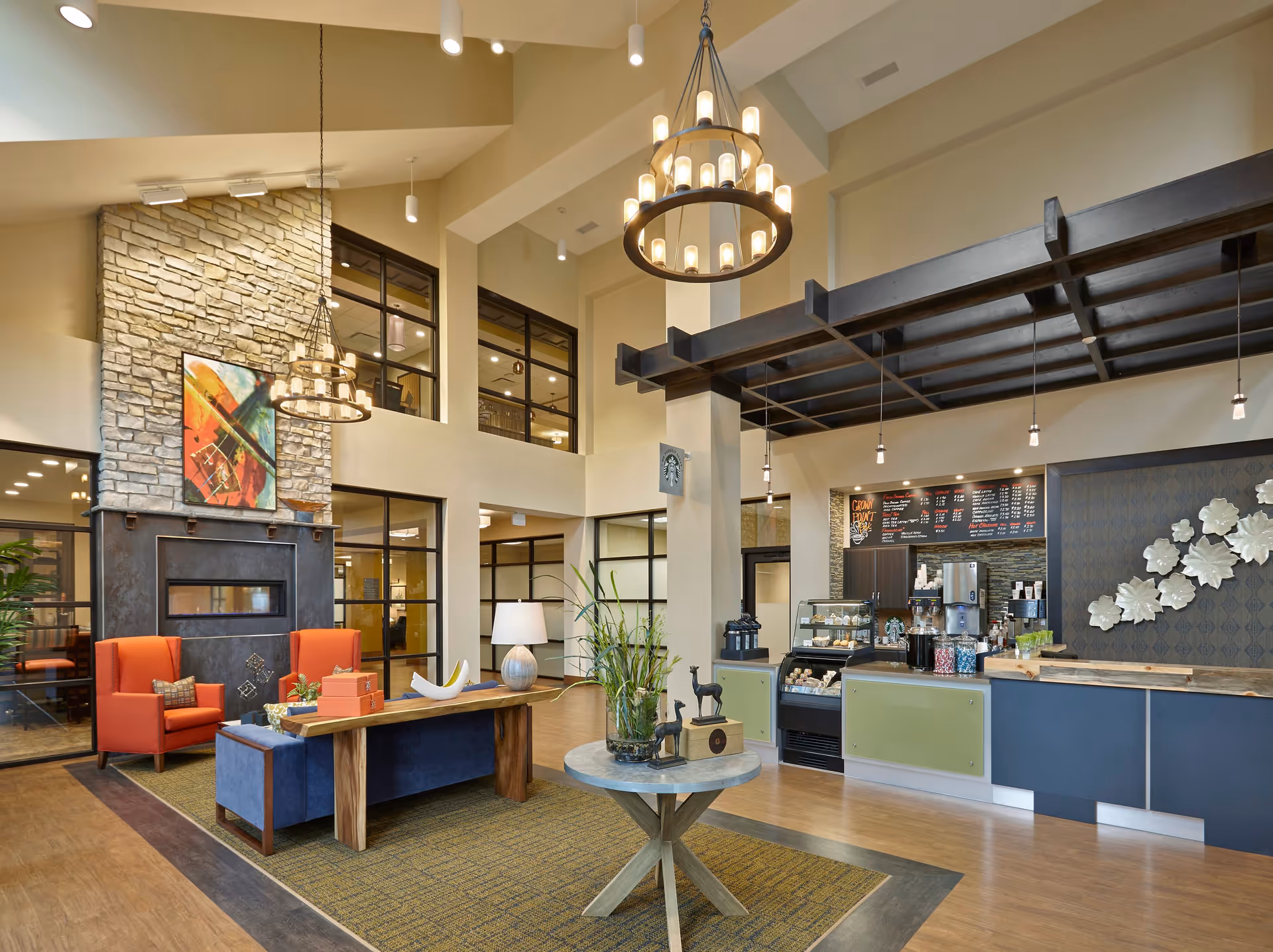 A spacious and well-lit interior lounge area of Ignite Medical Resorts Crownpoint featuring a modern coffee bar with a menu board, a stone fireplace with colorful artwork above it, comfortable seating including orange armchairs and a blue sofa, a round table with decorative items, and large windows allowing natural light.