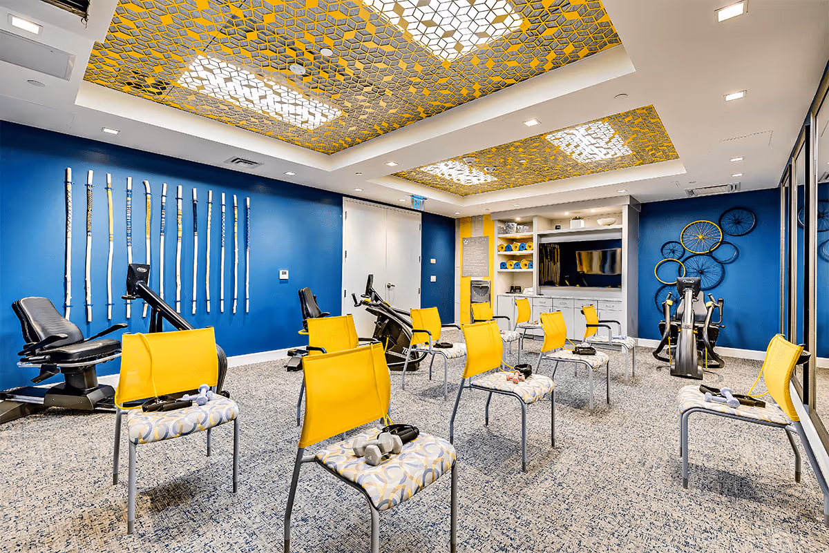 Bright exercise/activity room with yellow chairs arranged in rows, cardio equipment against blue walls, and decorative patterned ceiling panels.