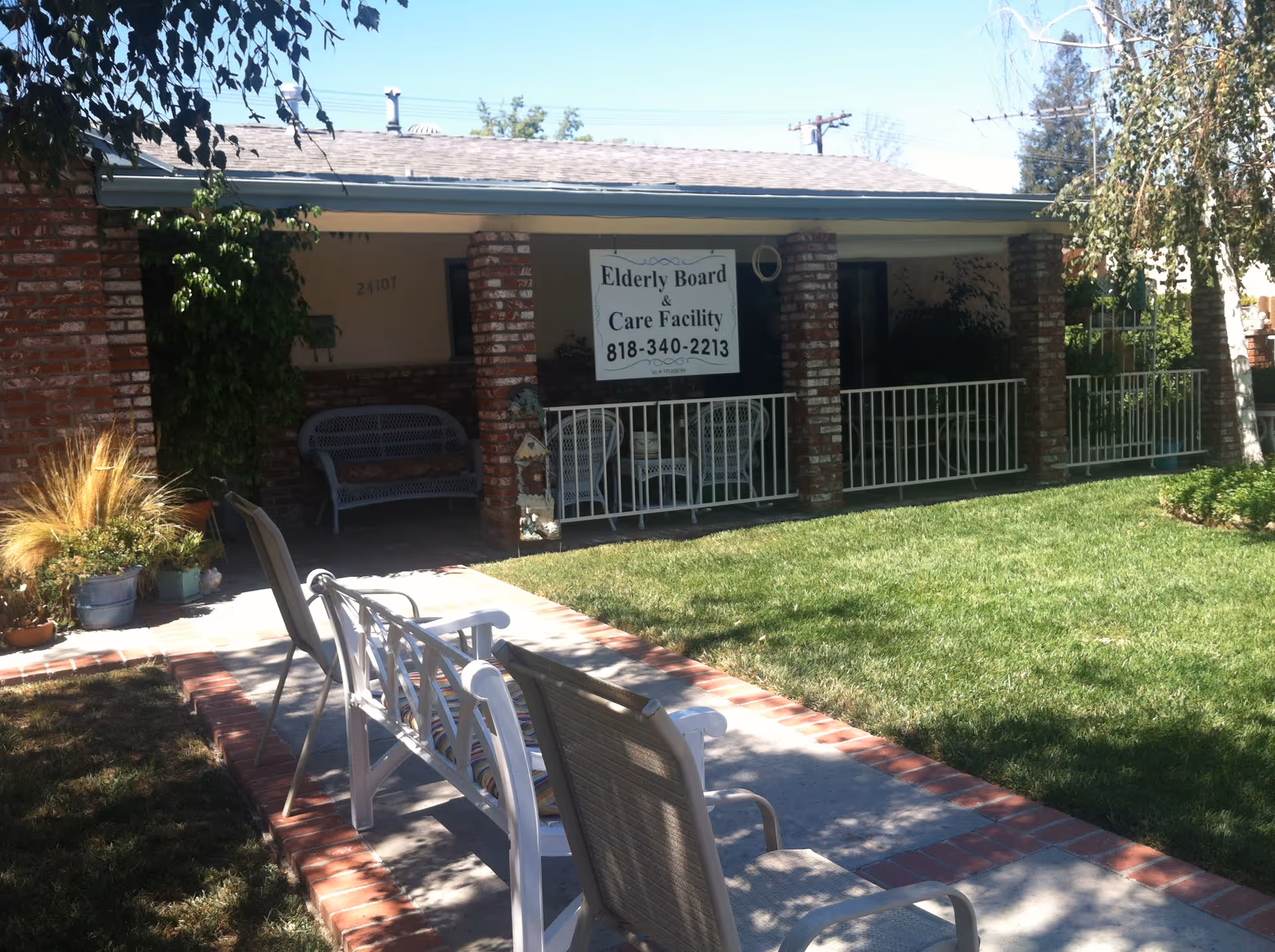 Front exterior view of a single-story brick building with a covered porch featuring white railings and outdoor seating. There is a green lawn and a paved walkway with chairs along it. A sign on the porch reads 'Elderly Board & Care Facility 818-340-2213'.
