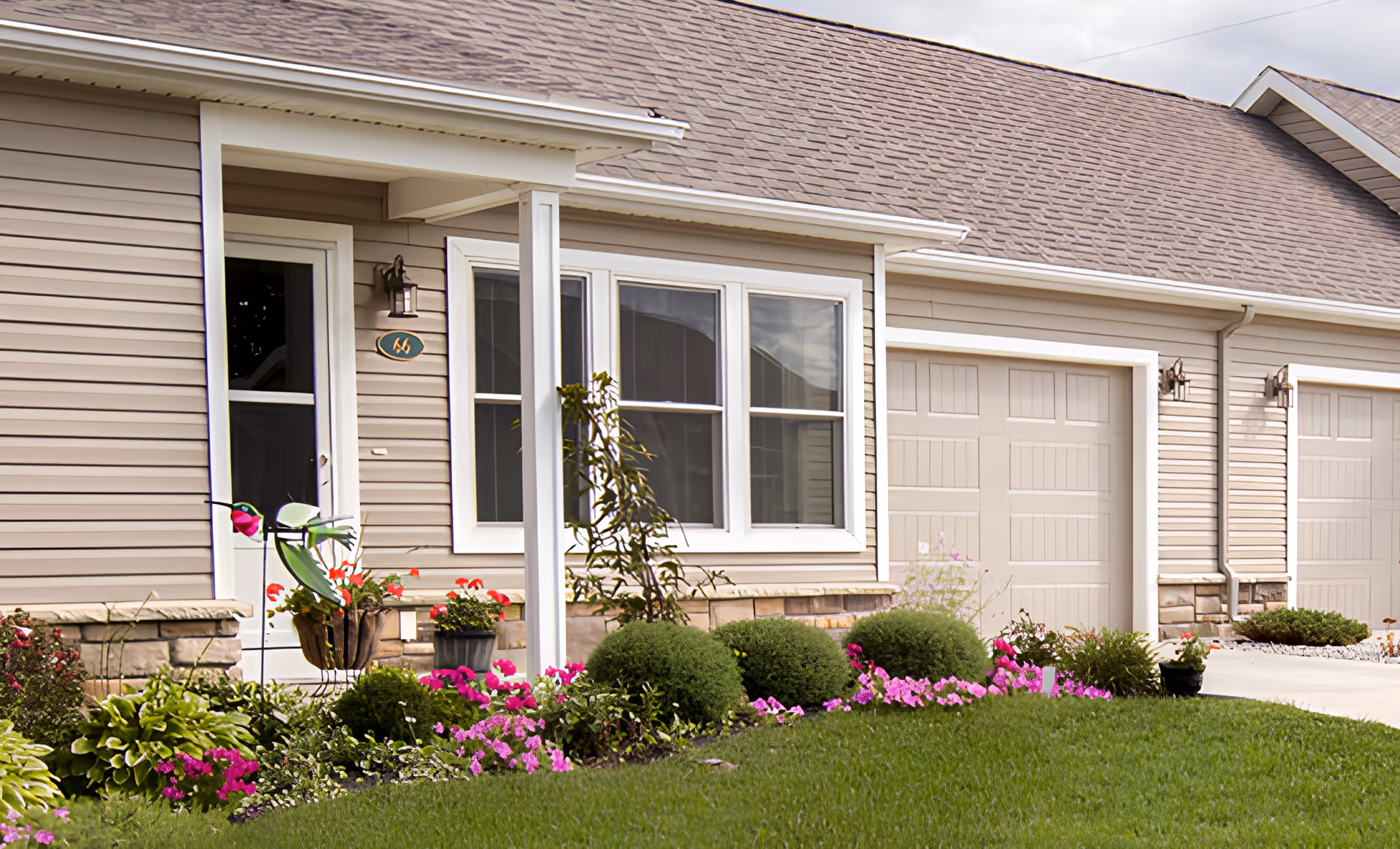 Exterior view of a senior living community unit with beige siding, a white-framed window, a white door, and a garage door. The front yard features a well-maintained lawn and a flower bed with green shrubs and pink flowers.