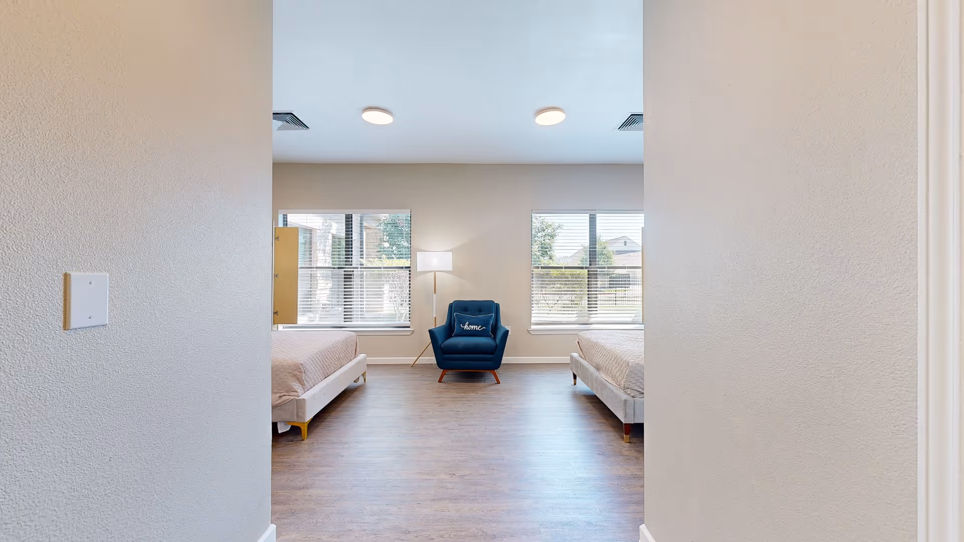 Entrance view into a bright shared bedroom with two beds, a blue armchair between windows and wood flooring.