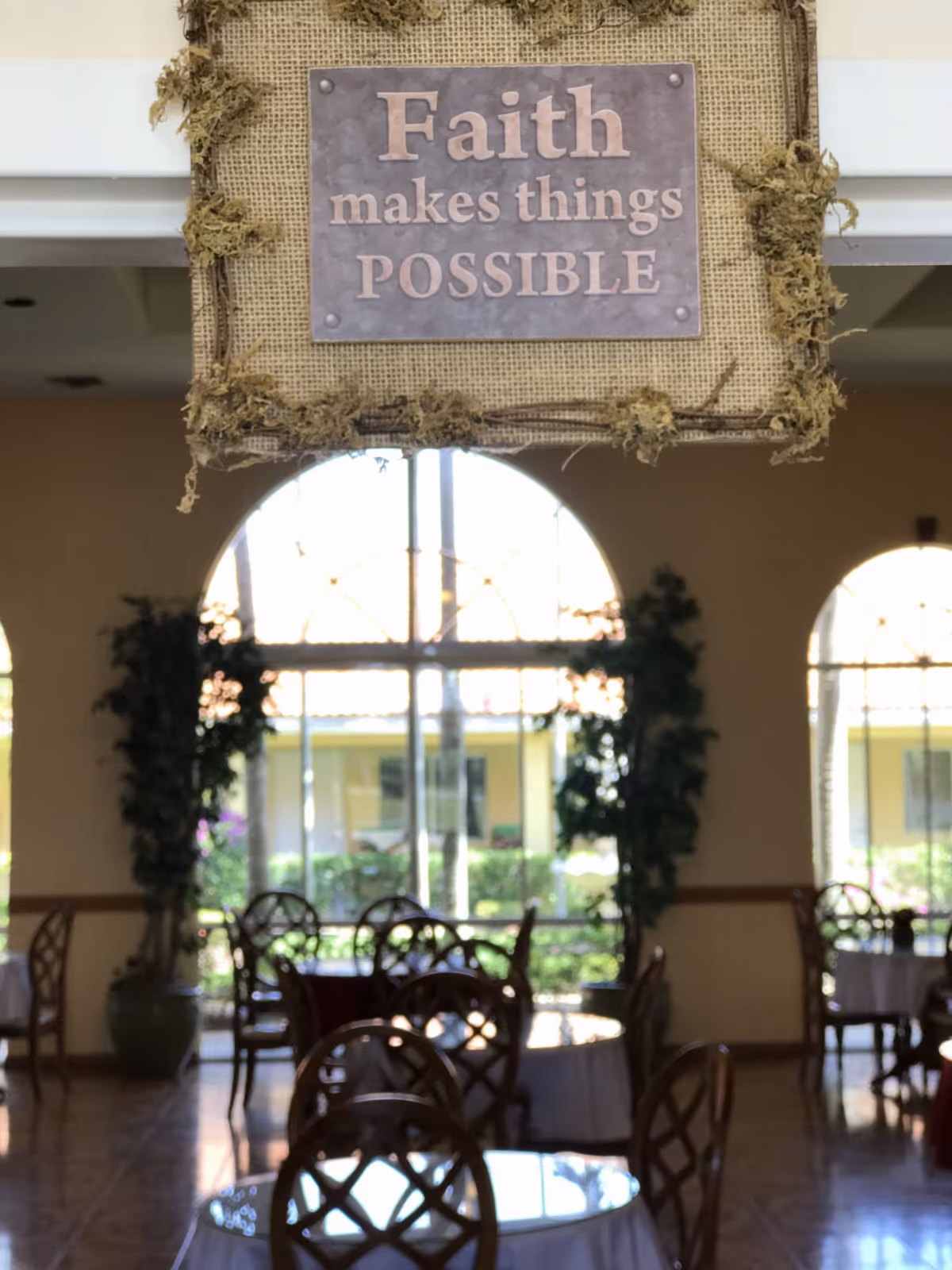 A decorative sign hanging indoors that reads 'Faith makes things POSSIBLE' with a dining area in the background featuring round tables with white tablecloths and wooden chairs. Large arched windows allow natural light to fill the room, and there are potted plants near the windows.